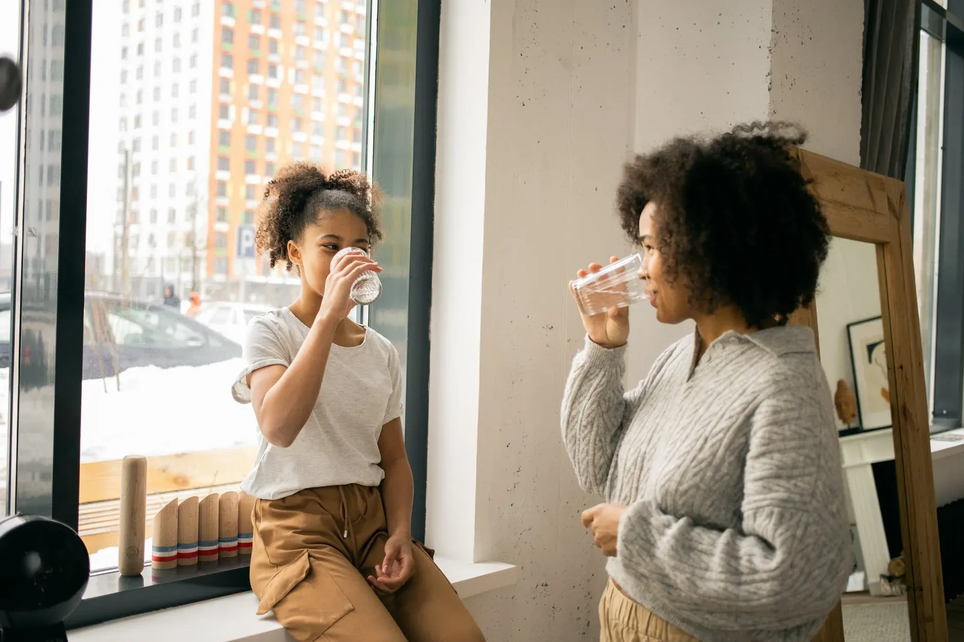 Imagem de uma mulher e uma menina segurando e bebendo um copo cheio de água olhando uma para a outra. Elas estão em ambiente interno e a menina está sentada em uma janela.