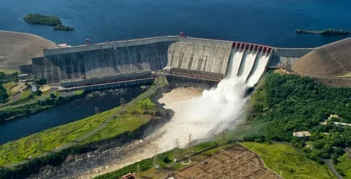 Uma das fontes de energias verdes é a hídrica. Mostrando isso, temos a imagem aérea de uma hidrelétrica, localizada em ambiente arborizado, com o mar ao fundo.