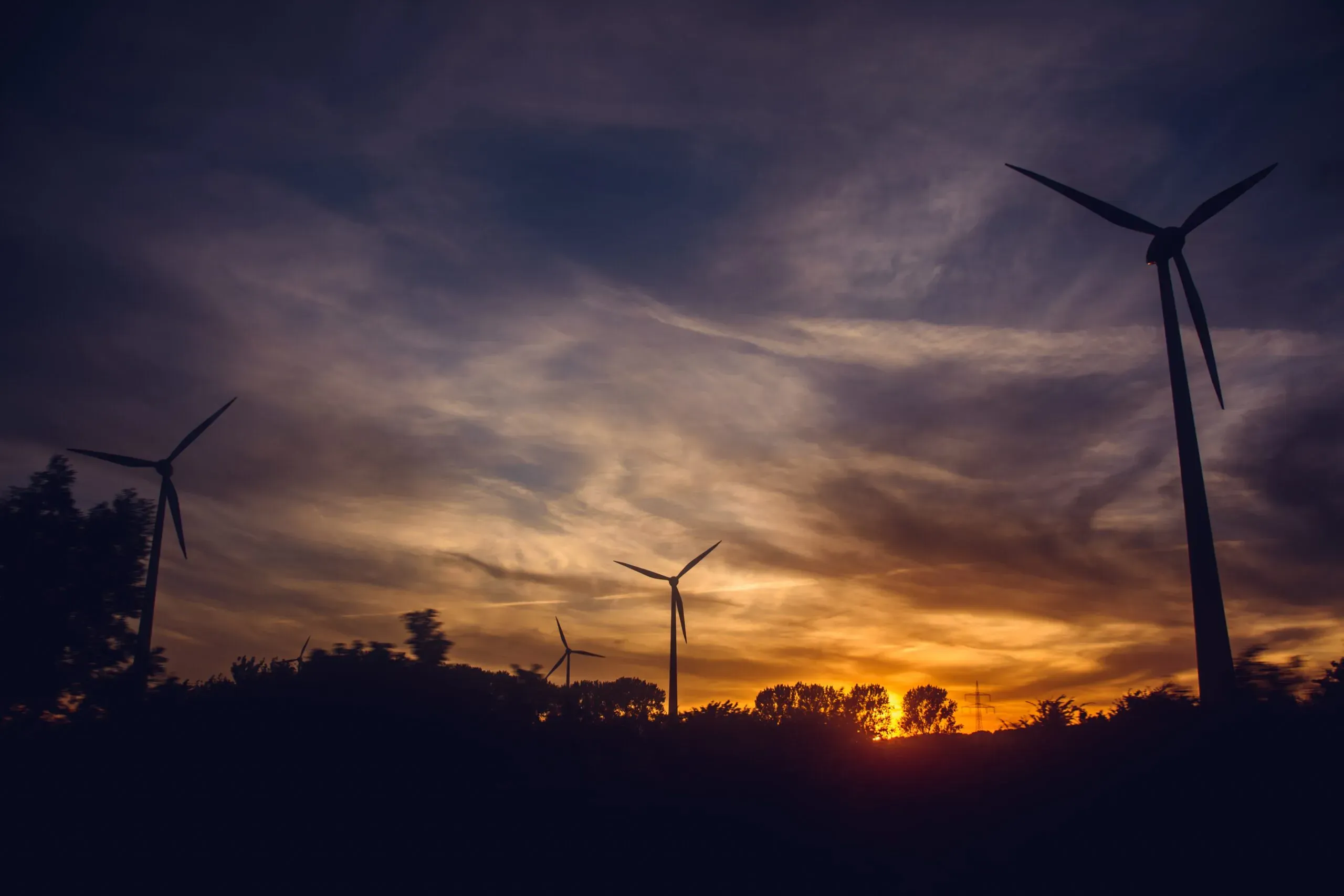 Em um ambiente ao ar livre, diante de um pôr do Sol, estão localizados moinhos de energia eólica.

