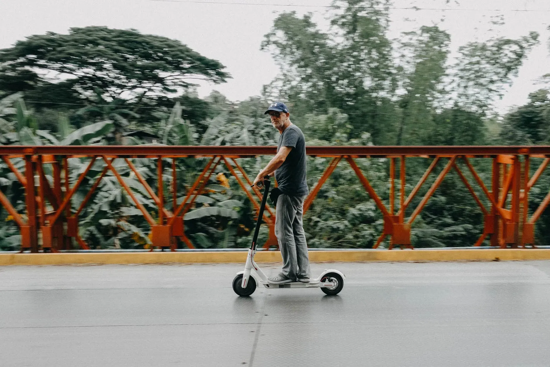 Imagem de um homem andando em um scooter elétrico. Ele veste boné, camiseta e calça jeans, e ao fundo há muitas árvores.