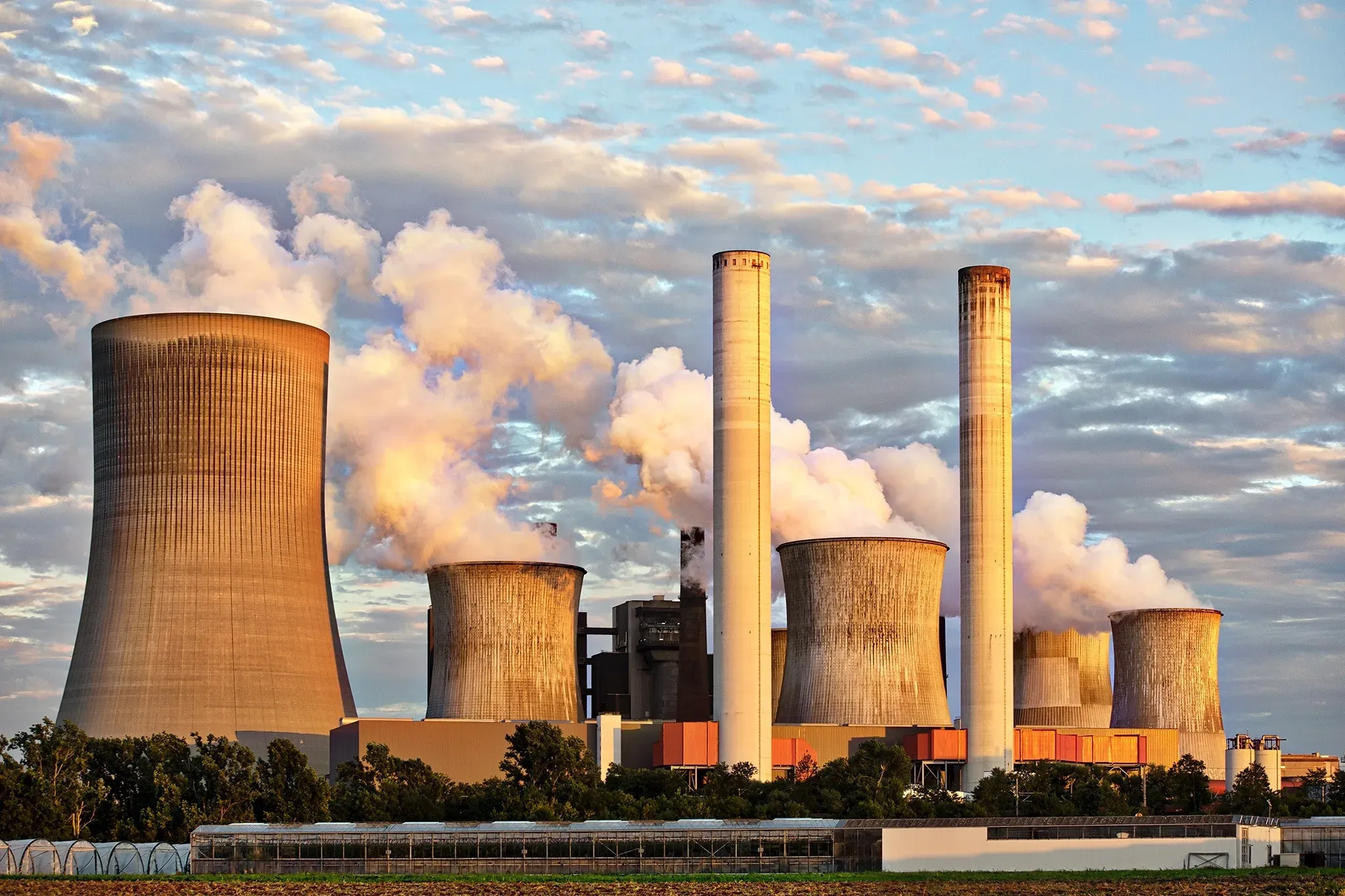 Imagem representando fontes de energia não renováveis. Uma usina nuclear é vista de dia. Há um prédio branco, com várias chaminés enormes soltando fumaça.
