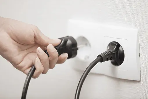 Em uma cozinha, uma mulher adulta se posiciona em frente ao fogão elétrico, apertando um botão, com luz natural vindo de uma grande janela ao fundo.